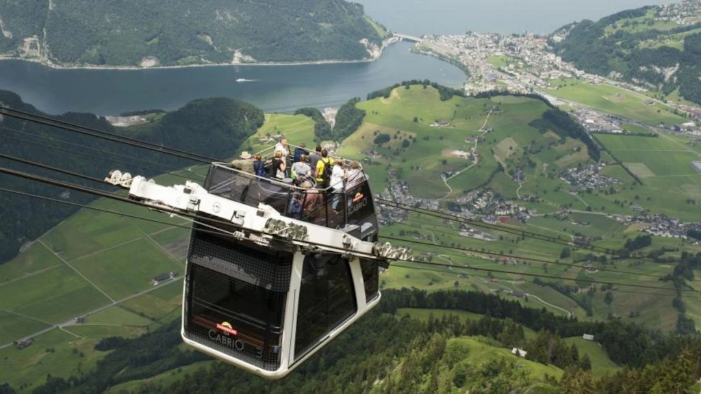Die CabriO-Seilbahn am Stanserhorn schwebt über einer grünen Landschaft, mit Passagieren an Bord, und überragt einen See sowie die im Tal verstreuten Dörfer.