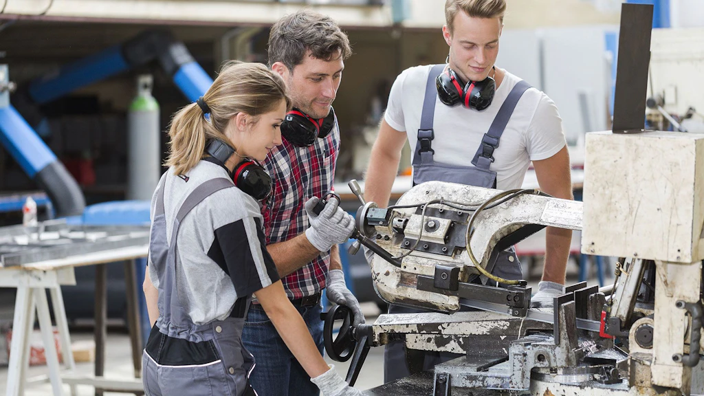 Ein Lehrmeister instruiert zwei Lernende bei der Bedienung einer Metallbearbeitungsmaschine.