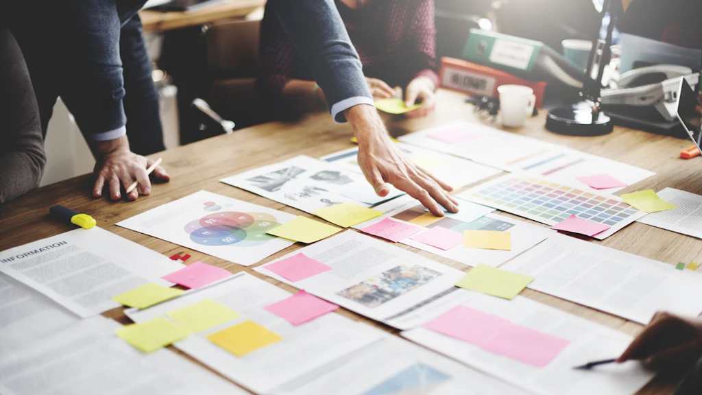 Diversi fogli annotati disposti su un tavolo, sui quali sono attaccati dei Post-it colorati, e mani e braccia visibili tutt'intorno al tavolo