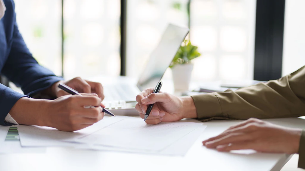 Deux personnes remplissent des formulaires sur table blanche, avec ordinateur portable, plante et fenêtre au fond.
