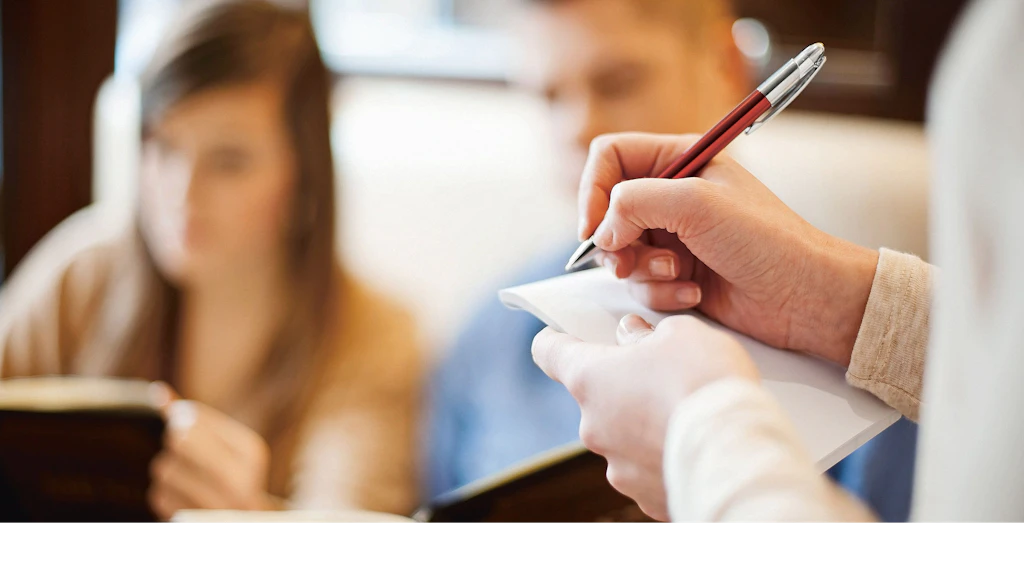 Person schreibt mit einem Stift Notizen auf einen Block, während im Hintergrund zwei unscharfe Personen sitzen