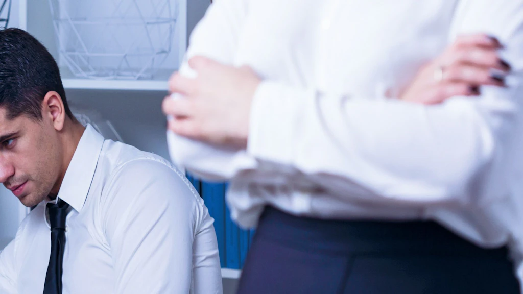 Nachdenklicher Mann im Büro sitzt am Schreibtisch, während im Vordergrund eine Person mit verschränkten Armen steht
