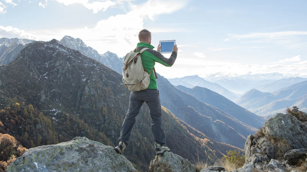A hiker stands on a rock and takes a photo with his iPad.