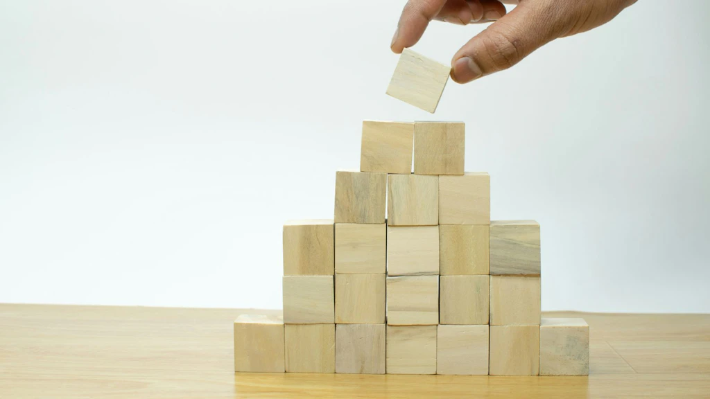 Zwei Finger einer Hand die eine Pyramide mit Holzklötzchen bauen