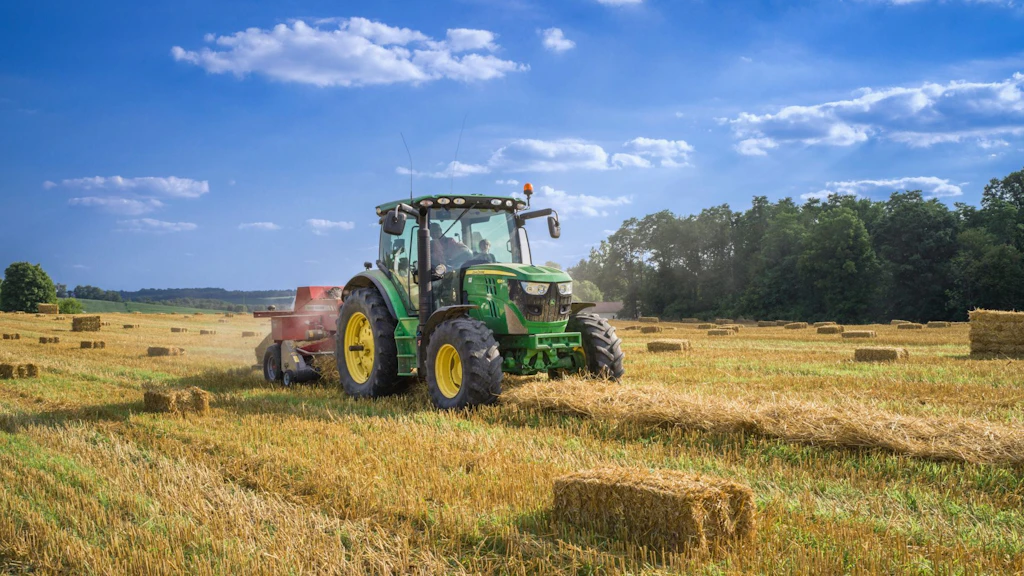 Grüner Traktor mit Ballenpresse auf einem Feld bei der Strohernte. Das Fahrzeug fährt über einen Strohschwad und presst rechteckige Strohballen
