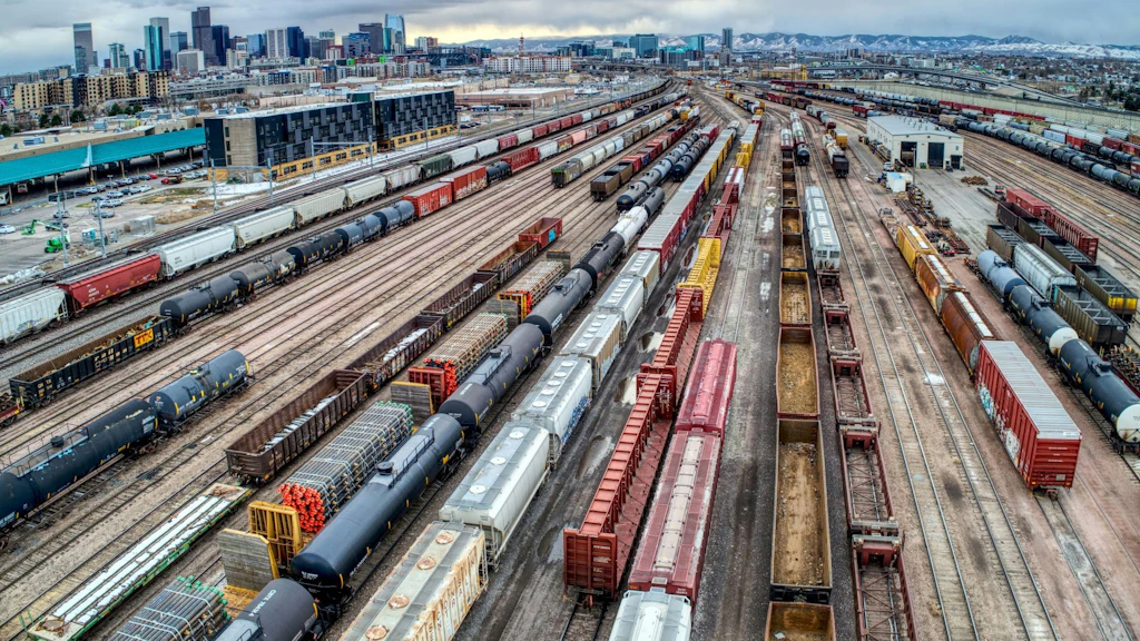 Großer Güterbahnhof mit zahlreichen parallel verlaufenden Gleisen und abgestellten Frachtzügen; im Hintergrund eine Stadt mit Hochhäusern und Bergen unter bewölktem Himmel