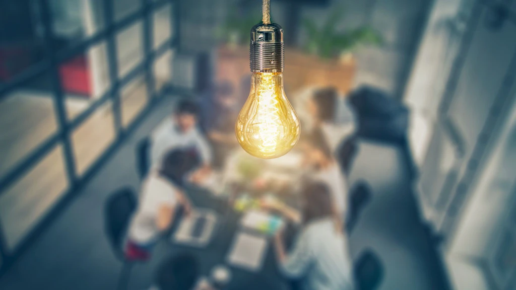 Leuchtende Glühbirne im Vordergrund, während im unscharfen Hintergrund ein Team an einem Tisch zusammenarbeitet und diskutiert.