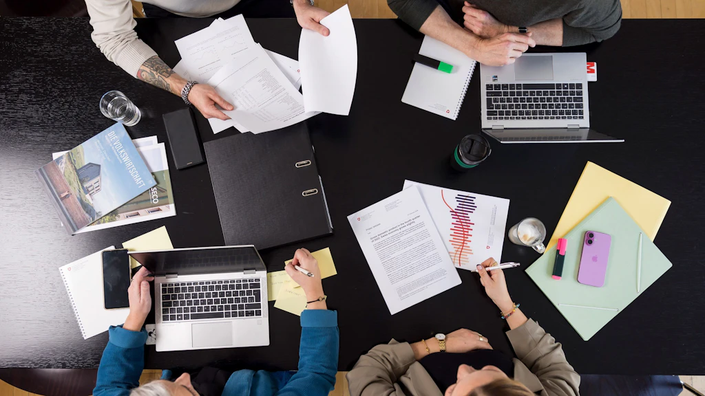 Konferenztisch – Szene aus einer gemeinsamen Arbeitssitzung – mehrere Personen arbeiten gemeinsam mit Laptops, ausgedruckten Unterlagen und Notizen. Auf dem Tisch liegen außerdem Ordner, ein Smartphone, Marker und Kaffeetassen.