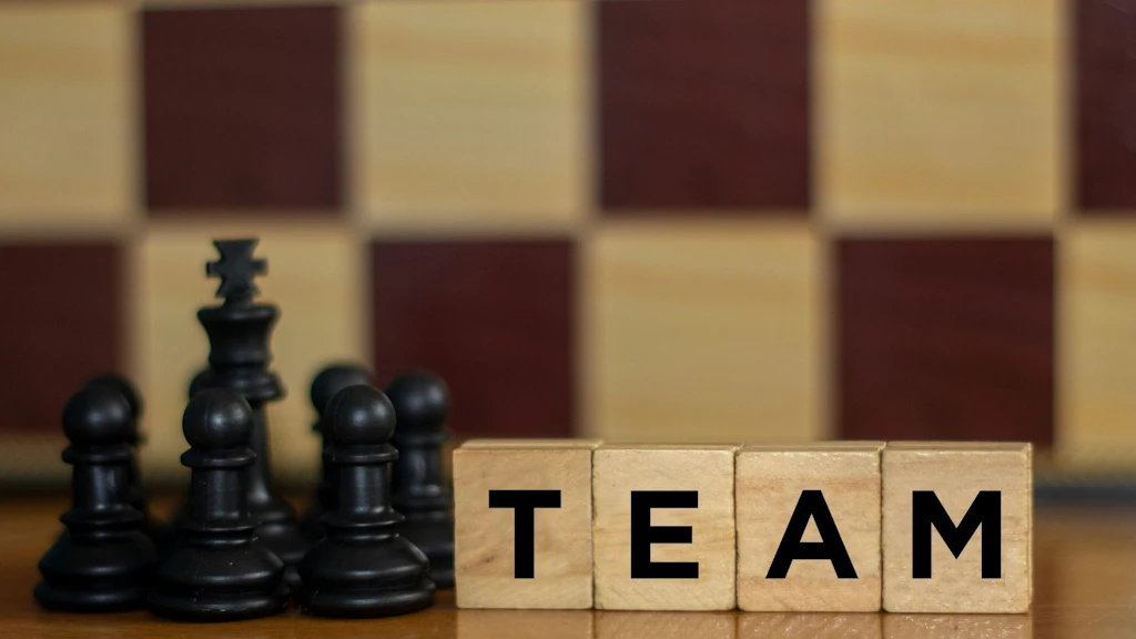 Schwarze Schachfiguren stehen vor einem Schachbrett. Im Vordergrund bilden vier Holzbuchstabenwürfel das Wort «TEAM». Das Bild symbolisiert Zusammenarbeit, Strategie und gemeinsames Handeln.