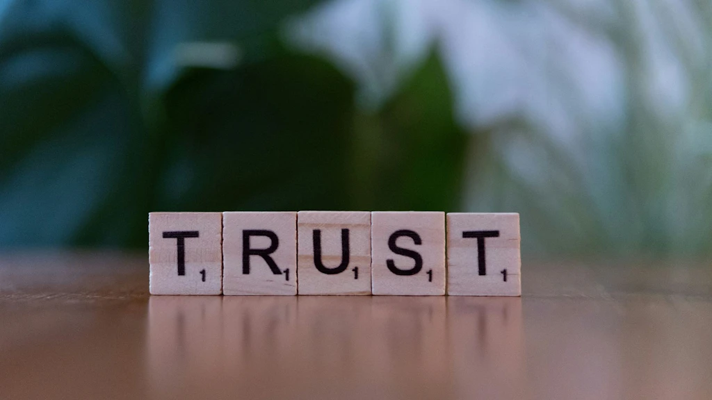 Five letter cubes on a table spell out the word "TRUST". The background is blurred and green. The image symbolizes trust, credibility, and reliability.