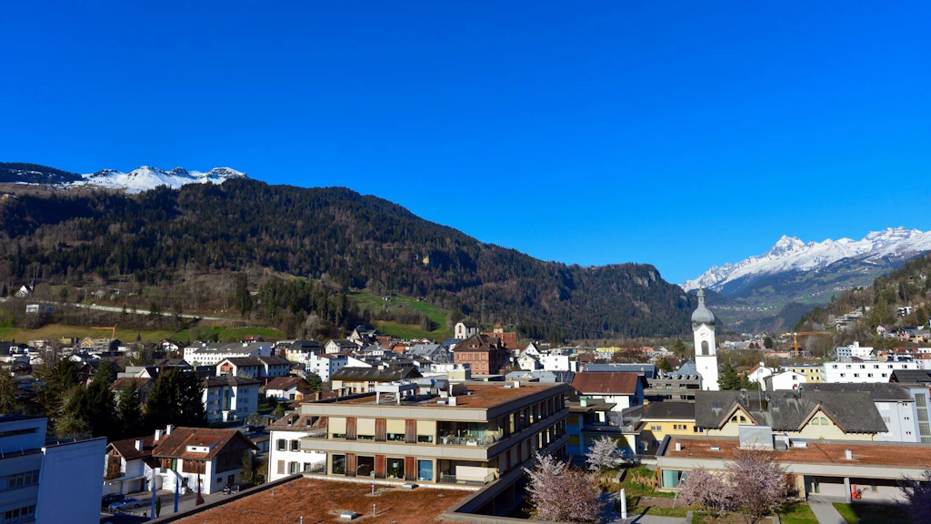 Panoramablick auf die Gemeinde Ilanz im Kanton Graubünden mit Häusern und einer Kirche mit Glockenturm, umgeben von bewaldeten Hügeln und schneebedeckten Bergen