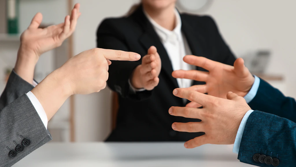Mehrere Personen in Businesskleidung gestikulieren mit den Händen während einer lebhaften Diskussion oder Verhandlung an einem Besprechungstisch.