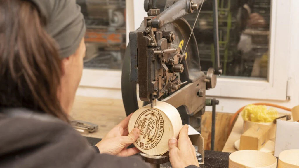 Eine Person bedient eine industrielle Maschine vom Typ mechanische Presse auf einer Holzwerkbank. Die Hände halten den Deckel des Vacherin Mont-d'Or-Käses, der mit einem dekorativen Muster verziert ist.