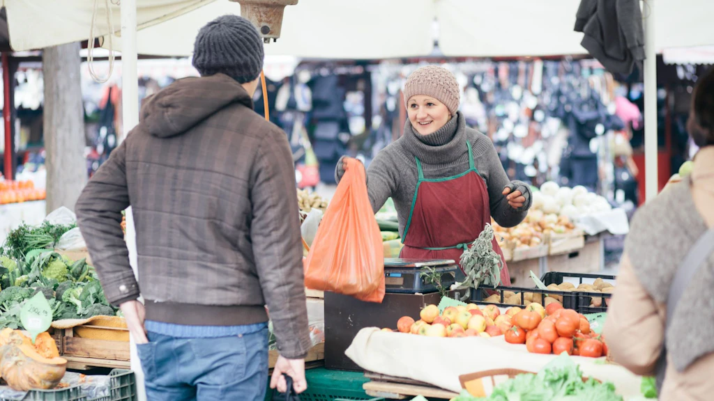 Eine Verkäuferin auf einem Markt im Freien reicht einem Kunden vor einem Stand voller frischem Obst und Gemüse eine Einkaufstüte.