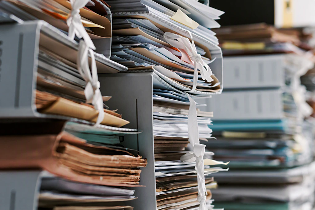 Nahaufnahme von überfüllten Regalen mit zahlreichen Papierakten, Mappen, Ordnern und Dokumentenstapeln. Die Unterlagen sind teilweise mit Schnüren zusammengebunden und übereinander gestapelt. Der Fokus liegt auf den Akten im Vordergrund, während der Hintergrund unscharf erscheint. Die Szene vermittelt den Eindruck eines großen Verwaltungs- und Dokumentationsaufwands sowie einer hohen Arbeitsbelastung durch papierbasierte Prozesse.
