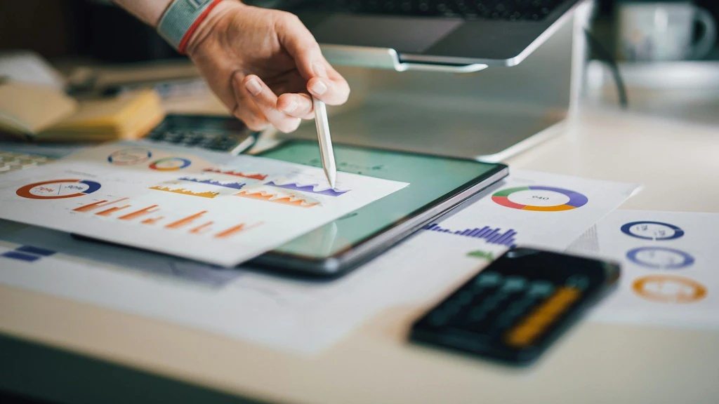 Eine Hand bedient mit einem Stift ein Tablet, auf dem bunte Grafiken zu sehen sind; daneben liegen Finanzunterlagen, ein Taschenrechner und ein Laptop auf einem Schreibtisch.