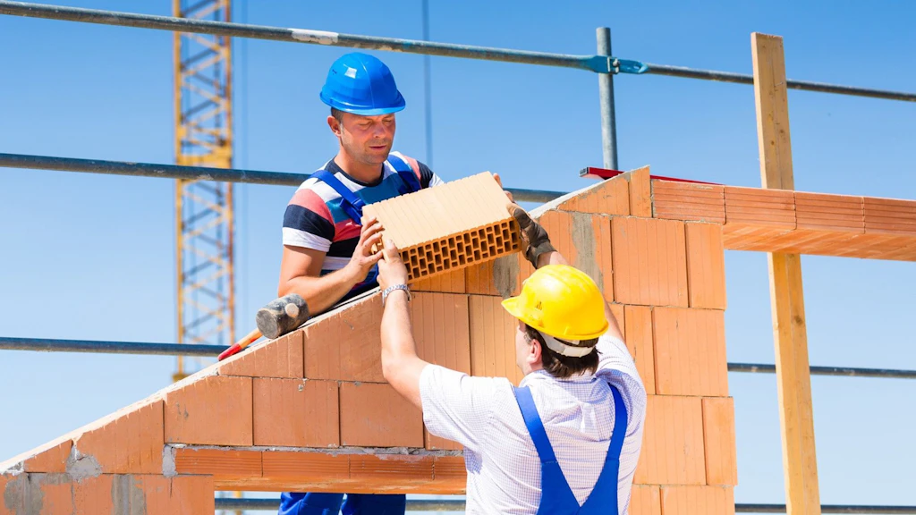 Zwei Bauarbeiter bauen eine Hausmauer und übergeben sich einen Ziegelstein.