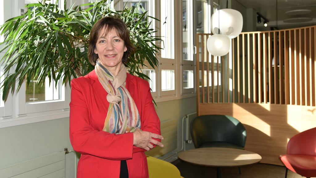 Frau in einem roten Blazer und einem bunten Schal steht neben einer Pflanze in einer Cafeteria.