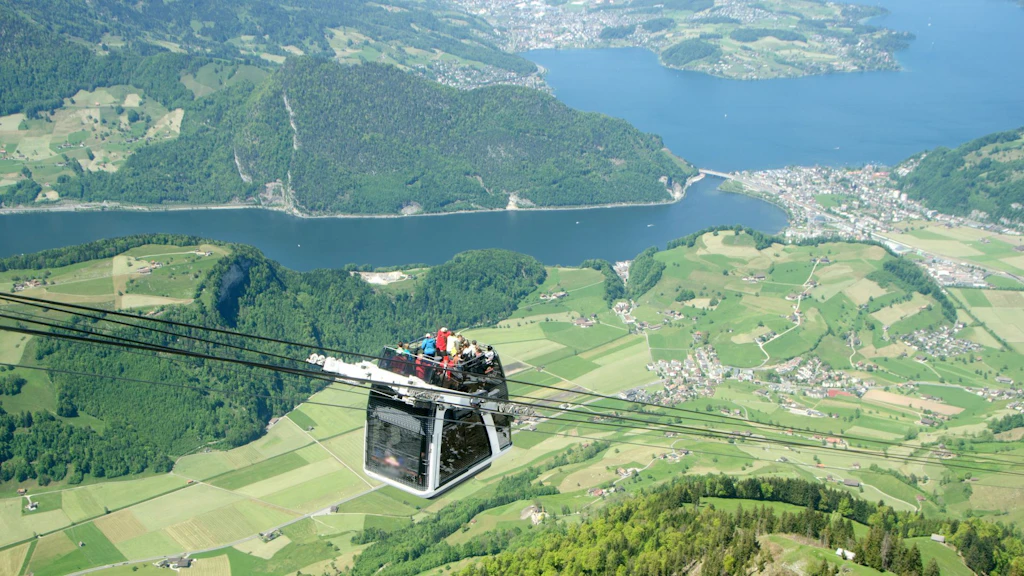 Die CabriO-Seilbahn am Stanserhorn schwebt über einer grünen Landschaft, mit Passagieren an Bord, und überragt einen See sowie die im Tal verstreuten Dörfer.