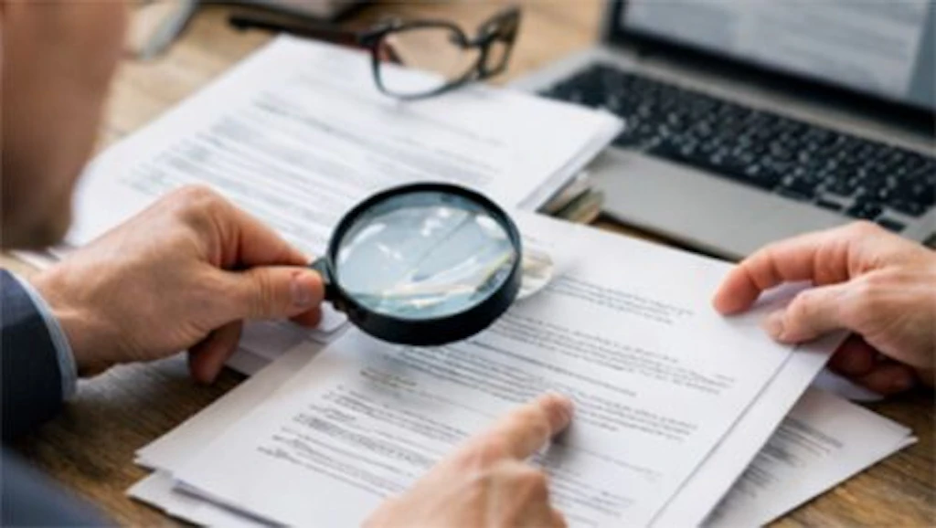 Table with documents, a person examining a document with a magnifying glass, a calculator, and a laptop.