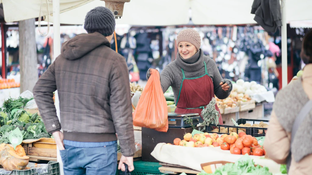 A market vendor hands a shopping bag to a customer in front of a stall laden with fresh fruit and vegetables.