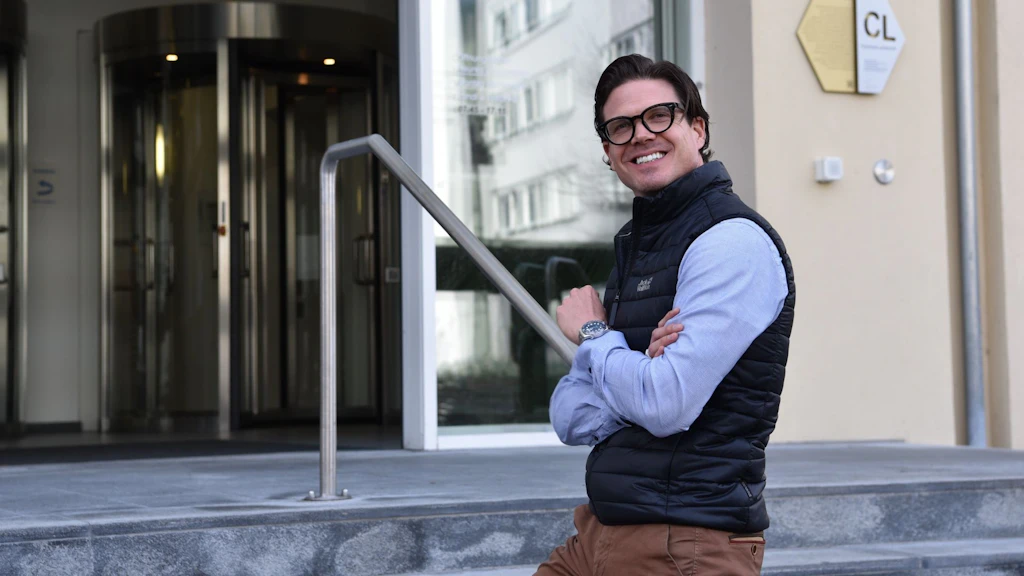 Homme portant des lunettes, un gilet noir et une chemise bleue, debout devant l'entrée du bâtiment du SECO.