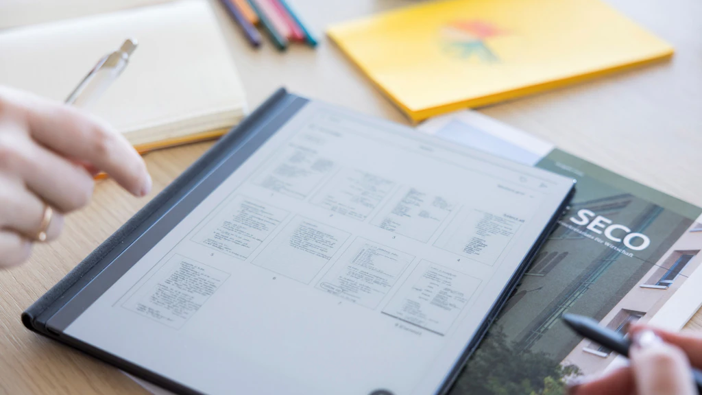 Deux mains tiennent un cahier ouvert contenant un tableau récapitulatif ainsi qu'un magazine du SECO, posés sur une table en bois clair sur laquelle se trouvent également des stylos, un bloc-notes et un cahier jaune.