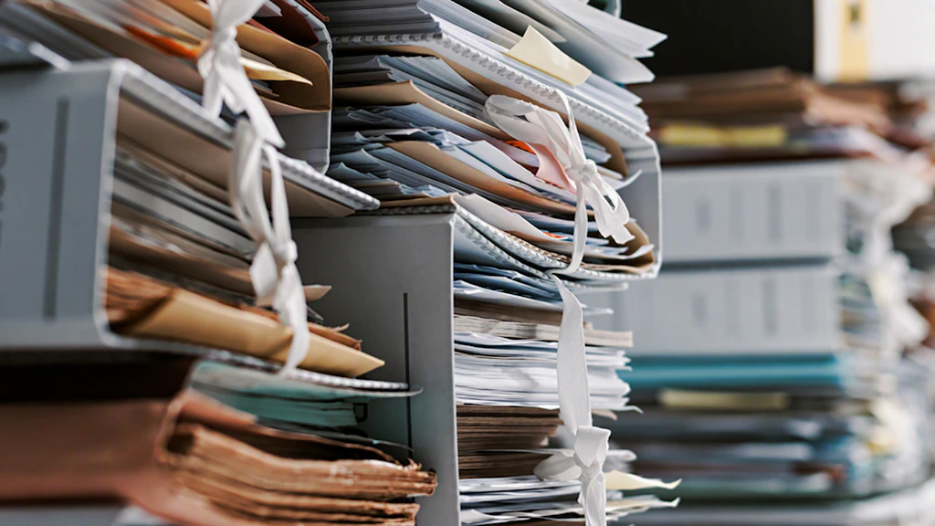 Gros plan sur des étagères débordantes de dossiers papier, de chemises, de classeurs et de piles de documents. Certains documents sont attachés avec de la ficelle et empilés les uns sur les autres. L'accent est mis sur les dossiers au premier plan, tandis que l'arrière-plan apparaît flou. La scène donne l'impression d'une charge administrative et documentaire importante, ainsi que d'une charge de travail élevée due aux processus papier.