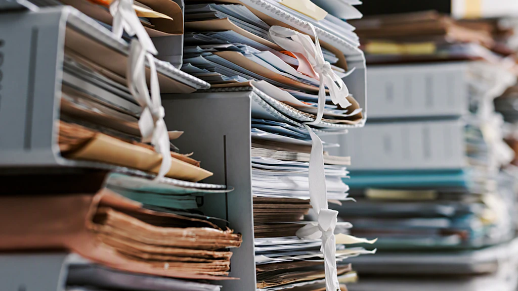 Gros plan sur des étagères débordantes de dossiers papier, de chemises, de classeurs et de piles de documents. Certains documents sont attachés avec de la ficelle et empilés les uns sur les autres. L'accent est mis sur les dossiers au premier plan, tandis que l'arrière-plan apparaît flou. La scène donne l'impression d'une charge administrative et documentaire importante, ainsi que d'une charge de travail élevée due aux processus papier.
