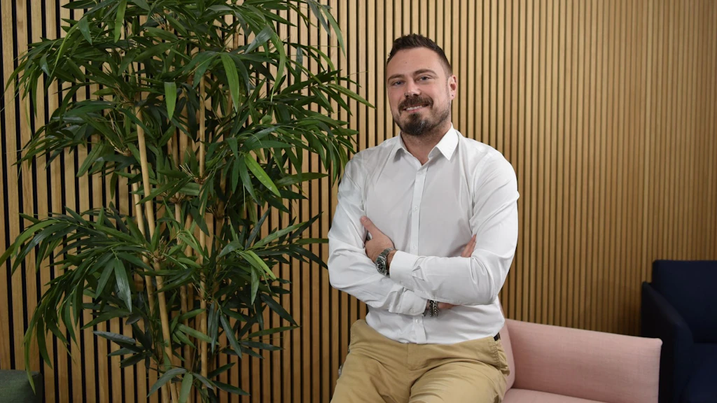 Homme barbu en chemise blanche assis devant un mur en bois avec une plante verte.