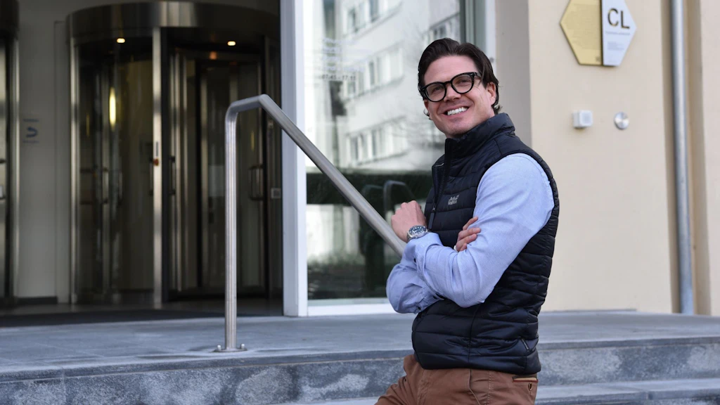 Uomo con occhiali, gilet nero e camicia blu in piedi davanti all'ingresso dell'edificio SECO