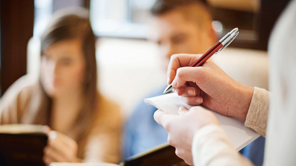 Una persona prende appunti con una penna su un blocco, mentre sullo sfondo sono sedute due persone sfocate